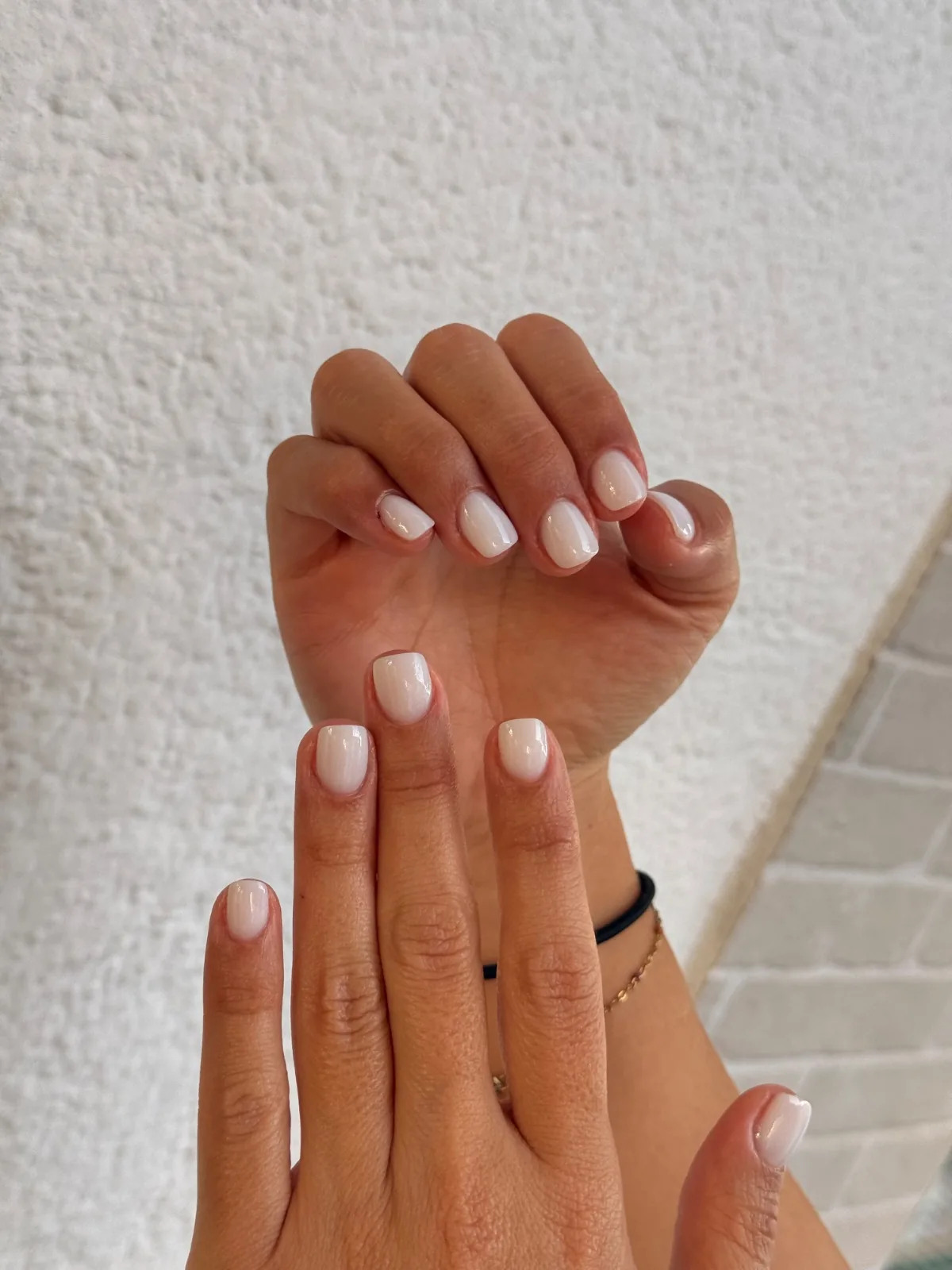 Manos con uñas cortas en tono lechoso natural sobre pared blanca texturada