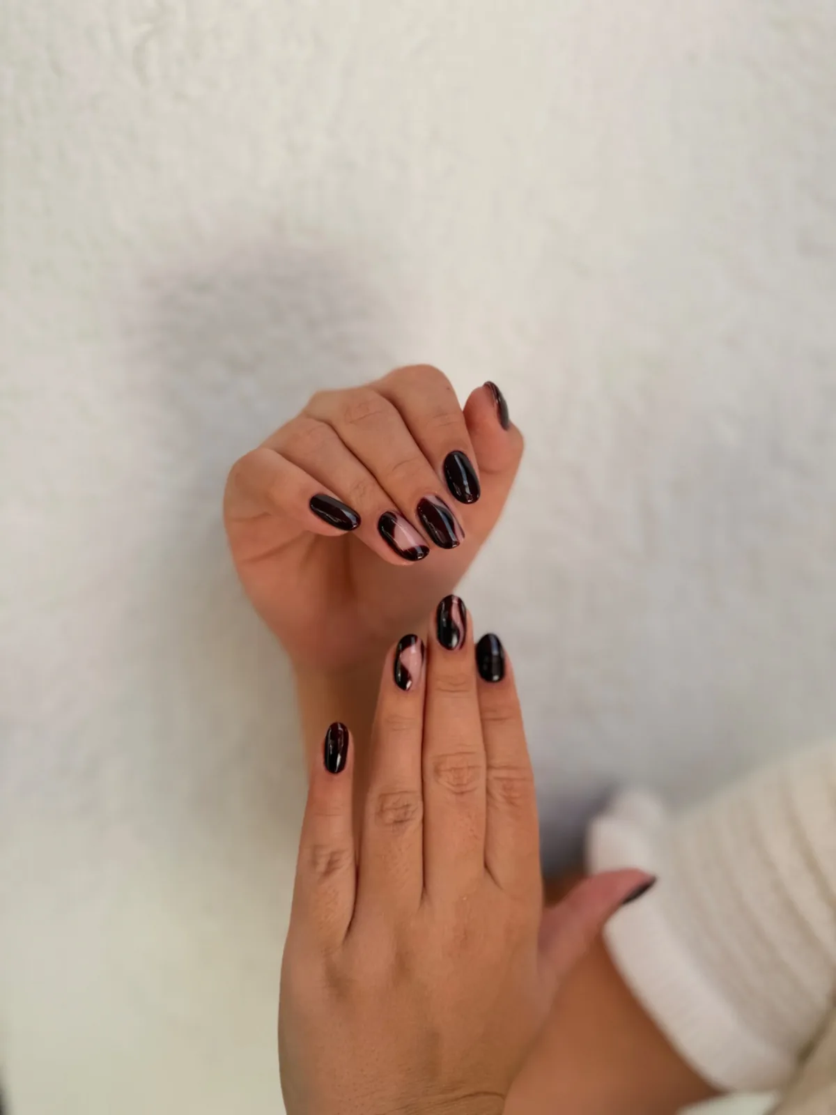 Manos con uñas ovaladas negras con diseño de transparencias rosadas sobre pared blanca